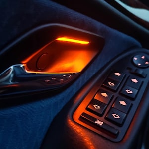 May include: A car interior door panel featuring a metallic silver door handle illuminated by a warm orange ambient light. Below the handle is a black control console with multiple rectangular buttons for window operation, each marked with directional arrows.