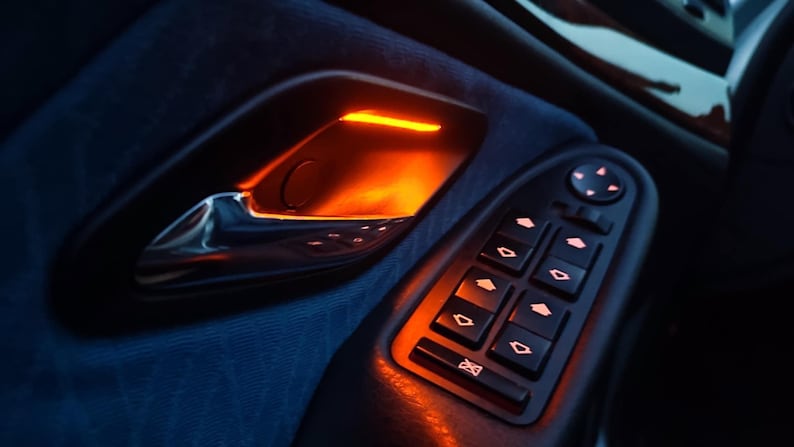 May include: A car interior door panel featuring a metallic silver door handle illuminated by a warm orange ambient light. Below the handle is a black control console with multiple rectangular buttons for window operation, each marked with directional arrows.