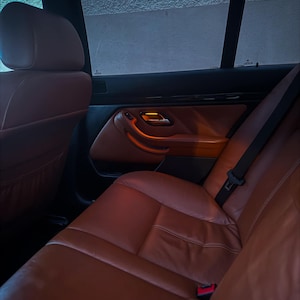 May include: The interior of a vehicle featuring cognac-colored leather seats and matching door panels. The cabin includes black trim accents, a seatbelt buckle, and a door handle with an illuminated orange light visible against the dark window.