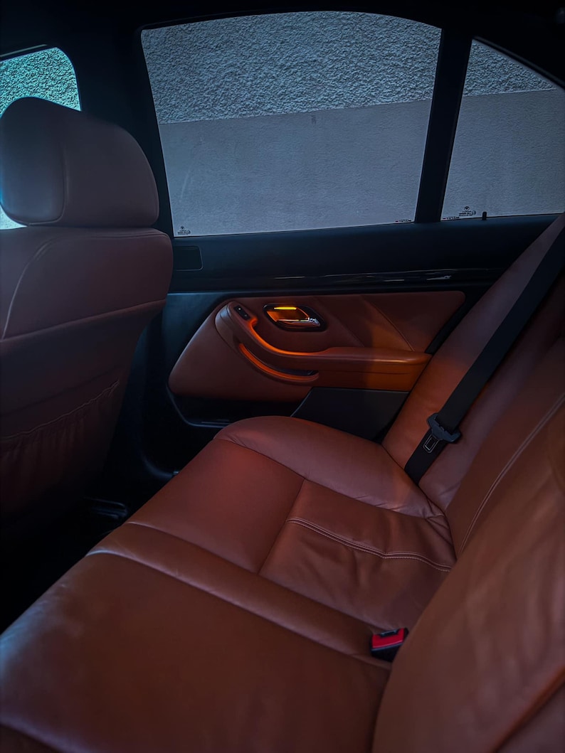 May include: The interior of a vehicle featuring cognac-colored leather seats and matching door panels. The cabin includes black trim accents, a seatbelt buckle, and a door handle with an illuminated orange light visible against the dark window.