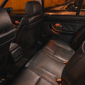 May include: The interior of a vehicle featuring black leather seats, headrests, and door panels. The cabin is illuminated by warm orange ambient lighting visible on the door handles and center console, with seat belt buckles visible on the bench.