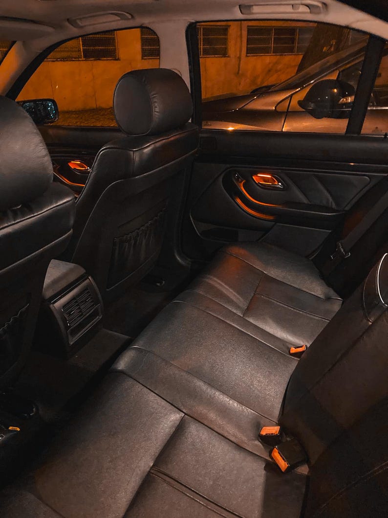 May include: The interior of a vehicle featuring black leather seats, headrests, and door panels. The cabin is illuminated by warm orange ambient lighting visible on the door handles and center console, with seat belt buckles visible on the bench.