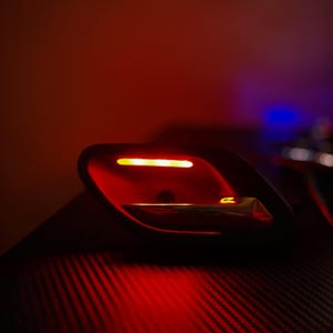 May include: A black automotive interior door handle assembly featuring a metallic silver lever and an integrated horizontal amber LED light strip, illuminated against a dark background with a subtle carbon fiber texture pattern.