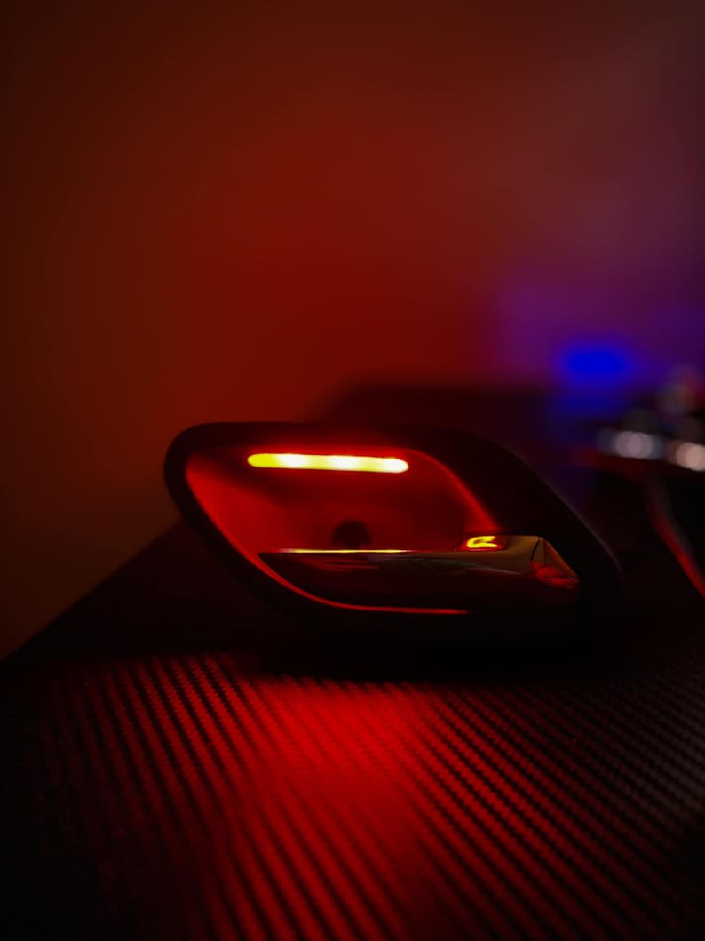 May include: A black automotive interior door handle assembly featuring a metallic silver lever and an integrated horizontal amber LED light strip, illuminated against a dark background with a subtle carbon fiber texture pattern.