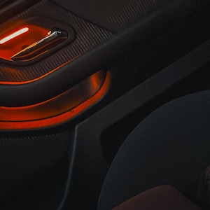 May include: A dark gray car door interior panel featuring a textured surface, a metallic door handle, and a recessed storage compartment illuminated by a bright orange ambient light strip that highlights the contours of the door panel design.