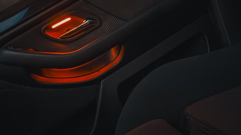 May include: A dark gray car door interior panel featuring a textured surface, a metallic door handle, and a recessed storage compartment illuminated by a bright orange ambient light strip that highlights the contours of the door panel design.