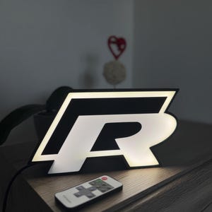 Könnte beinhalten: Ein schwarz-weiß beleuchtetes Schild in Form des Buchstabens „R“. Das Schild wird von einem hellen weißen Licht beleuchtet und steht auf einer Holzoberfläche. Eine Fernbedienung befindet sich vor dem Schild.