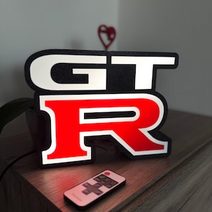 Op de afbeelding: Een verlicht bord met de letters "GT-R" in wit en rood, met een zwarte rand. Een afstandsbediening ligt voor het bord. Het bord staat op een houten oppervlak.