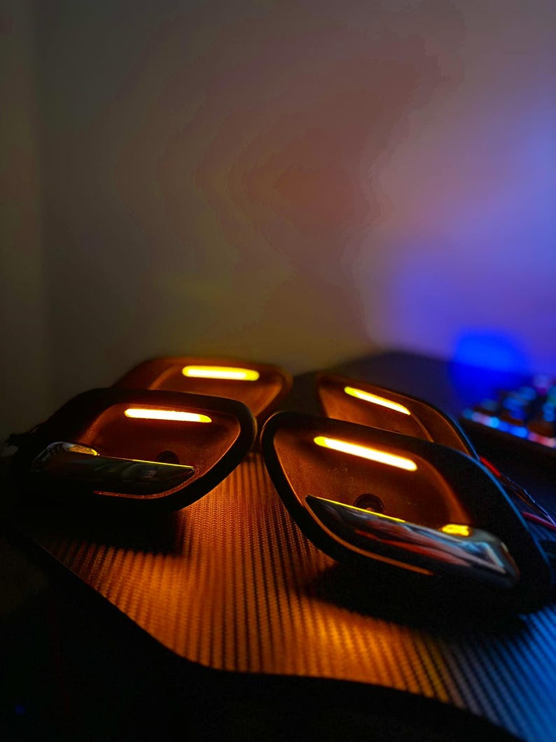 May include: A set of four black automotive interior door handle assemblies featuring integrated amber LED light strips and polished chrome levers. Each unit has a contoured housing with a horizontal light bar positioned above the handle.