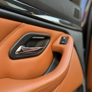 May include: A car door interior panel featuring tan leather upholstery with visible stitching, a matte black door handle housing, a polished chrome interior door release lever, and a small black control button panel integrated into the armrest.