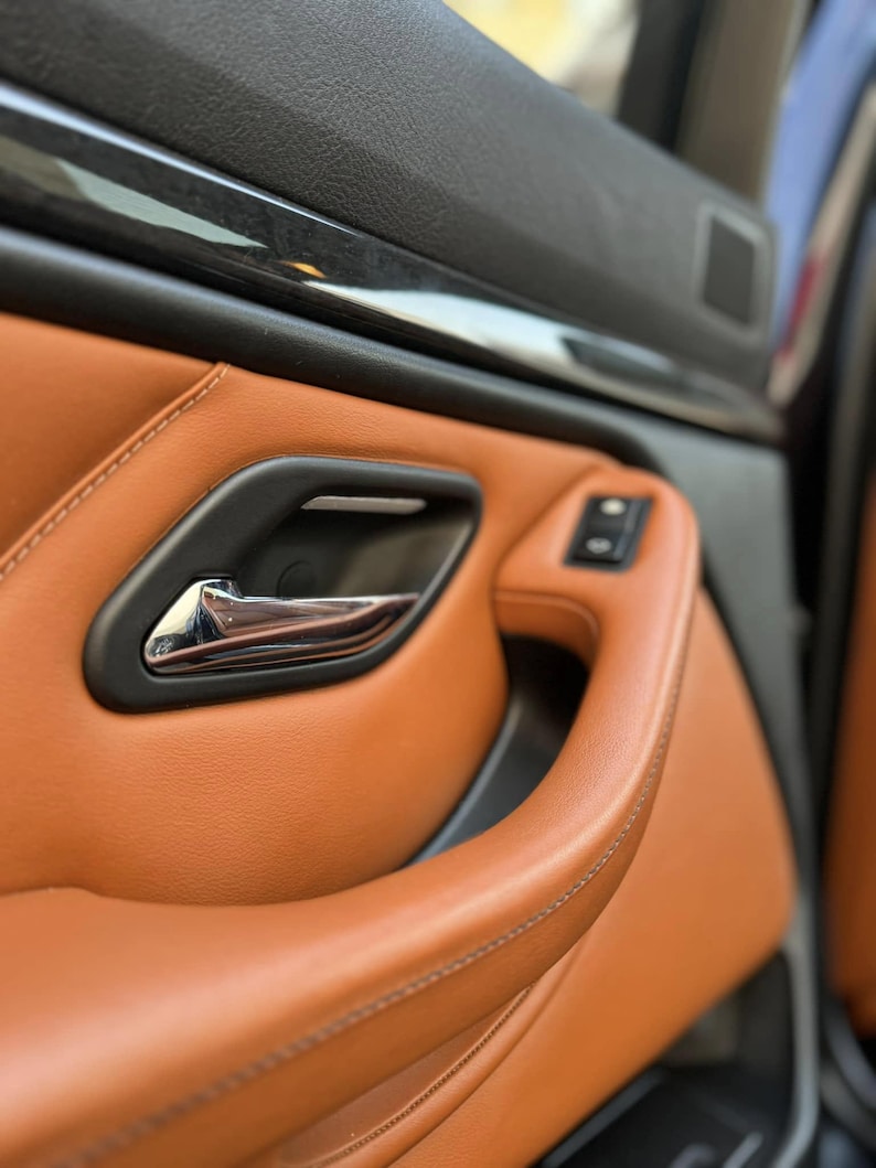 May include: A car door interior panel featuring tan leather upholstery with visible stitching, a matte black door handle housing, a polished chrome interior door release lever, and a small black control button panel integrated into the armrest.