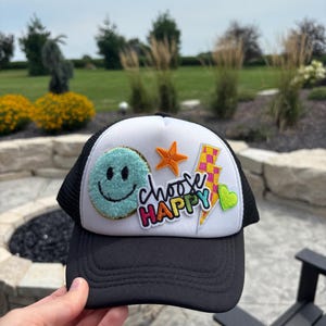 Puede incluir: Gorra de camionero en blanco y negro con un panel frontal blanco con parches. Los parches incluyen una carita sonriente turquesa, una estrella naranja, un rayo, un corazón verde y las palabras "choose HAPPY".