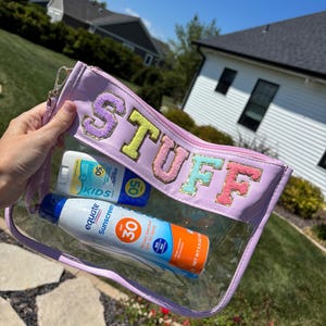 Puede incluir: Un neceser rectangular transparente con ribete lavanda y la palabra "STUFF" en letras bordadas de colores. En el interior hay dos botellas de protector solar, una para niños con SPF 50 y otra con SPF 30.