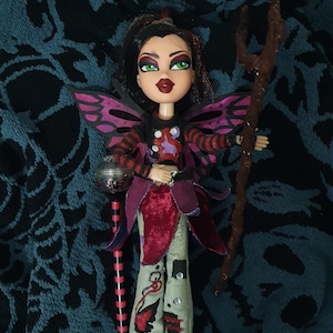 May include: A doll with green eyes, dark hair, and red lips, wearing a black and red top, a burgundy and purple outfit, and gray pants with red and black designs. The doll has pink and purple wings and holds a staff and a decorative scepter.