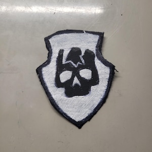 May include: A shield-shaped patch with a black skull design on a white background. The skull has a cracked appearance. The patch is outlined in black and appears to be made of fabric.