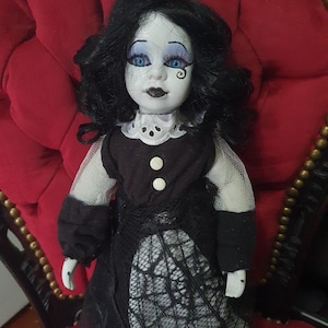 May include: A porcelain doll with black curly hair, blue eyes, and dark lipstick. The doll is dressed in a black dress with a white lace overlay and a white collar. The doll is seated on a red velvet chair.
