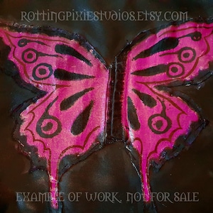 Puede incluir: Una mariposa rosa con marcas negras está pintada sobre una superficie de cuero negro. La mariposa tiene un cuerpo central grande y dos alas con detalles intrincados. El texto "EXAMPLE OF WORK, NOT FOR SALE" está impreso debajo de la mariposa.