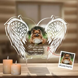 May include: A clear acrylic heart-shaped memorial with angel wings, a photo frame, and the text "God has you in HIS ARMS I have you in my Heart".