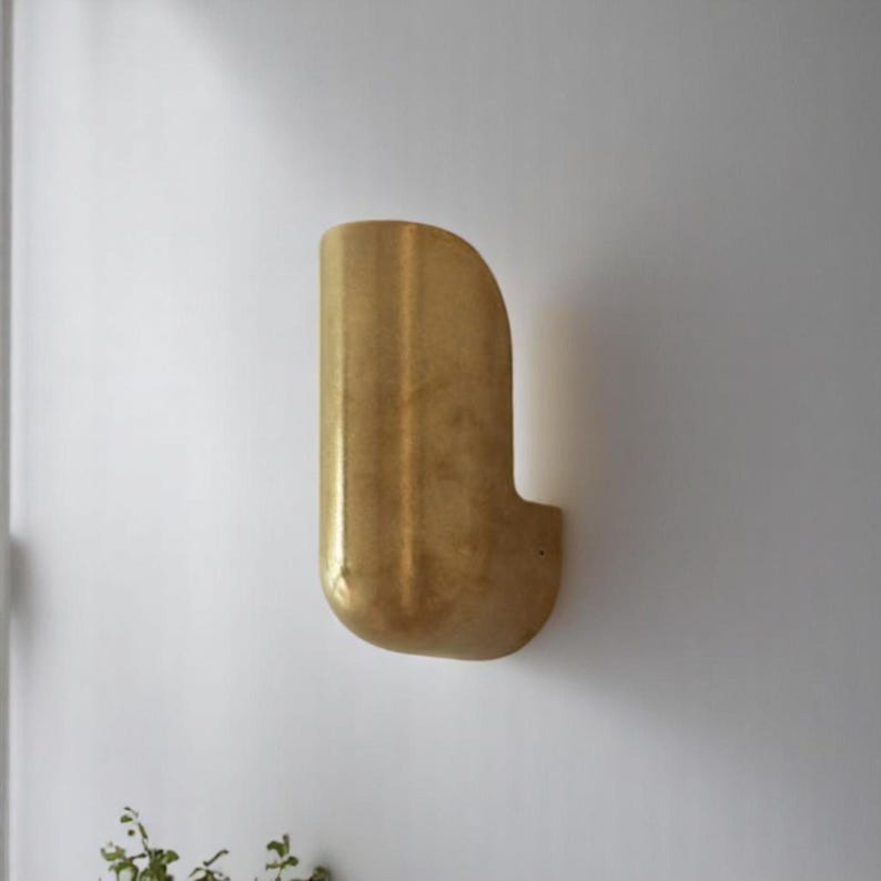 May include: A gold-colored wall sconce with a curved, abstract design. The sconce is mounted on a white wall.
