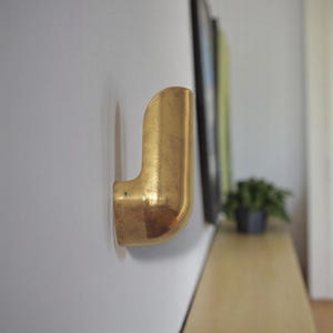 May include: A gold-colored metal hook mounted on a white wall. The hook is shaped like a curved pipe.