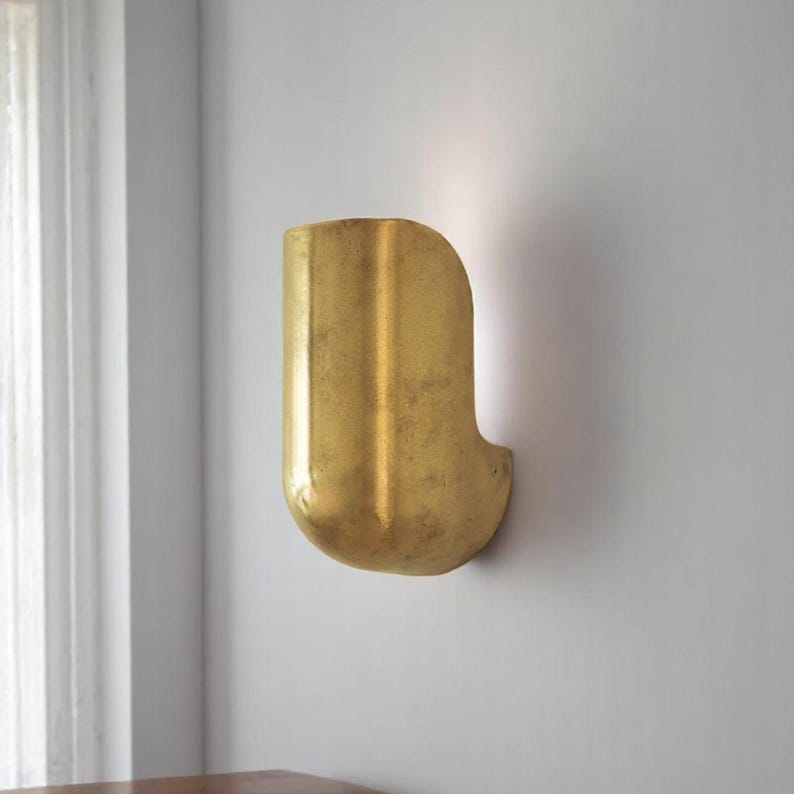 May include: A gold-colored wall sconce with a curved, rectangular shape. The sconce has a textured surface and is mounted on a white wall.