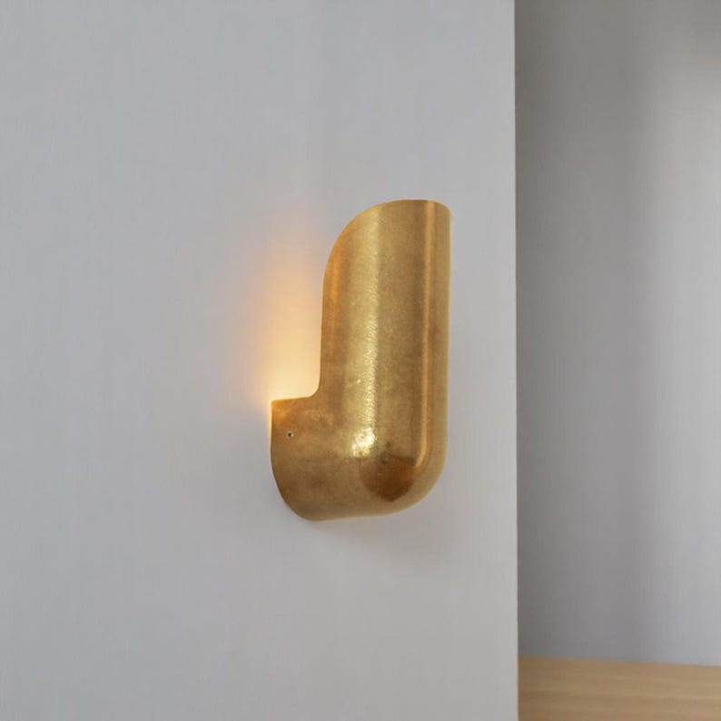 May include: A gold-toned wall sconce with a curved, cylindrical design. The sconce is mounted on a white wall and emits a warm, ambient light.