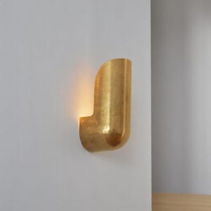 May include: A gold-toned wall sconce with a curved, cylindrical design. The sconce is mounted on a white wall and emits a warm, ambient light.