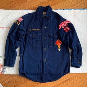 May include: A navy blue Cub Scout uniform shirt with a patch that says "CUB SCOUTS B.S.A." and a patch with the text "80 ST. PAUL MINN. 79".