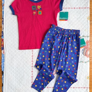 May include: A children's pajama set featuring a pink short-sleeved top with blue trim and embroidered squares. The blue pajama pants have a pattern of yellow, red, and blue shapes. The set is displayed on a white quilted surface.