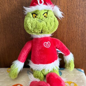 May include: A plush Grinch doll wearing a red Santa hat and a red outfit with white trim. The Grinch has green fur, yellow eyes, and a white beard. A white heart is on the chest of the red outfit. The doll is sitting on a patterned surface.