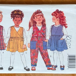 May include: Butterick sewing pattern envelope featuring illustrations of four young girls in various outfits. The outfits include a plaid dress, a wrap-style dress, floral overalls, and a blue dress. The envelope is white with the words "FAST & EASY" and "Butterick".