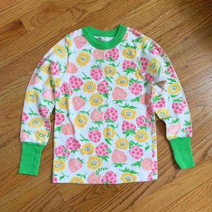 May include: A white long-sleeved shirt with a green collar and cuffs. The shirt has a repeating pattern of pink strawberries and yellow flowers.