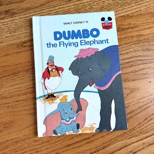 May include: A vintage Walt Disney's "Dumbo the Flying Elephant" book. The cover features illustrations of Dumbo, a circus ringmaster, and other characters. The book has a light blue cover with the title in large blue letters.