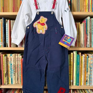 May include: A vintage children's outfit featuring a white long-sleeved shirt and navy blue overalls with red suspenders. The overalls have a Winnie the Pooh patch and a red "P" on the leg. A "Pooh" tag is attached.