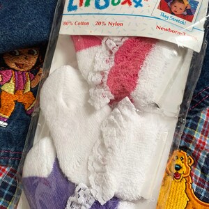 May include: A package of three pairs of white baby socks with lace trim. The socks are pink, white, and purple. The package is labeled "Lil Soxx" and says "For Newborns" and "They Stretch!"