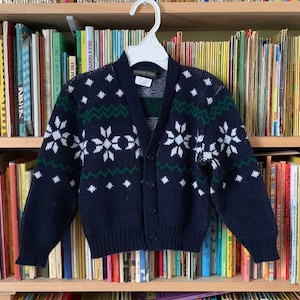 May include: A navy blue cardigan sweater with a white and green snowflake pattern. The cardigan has a V-neck and button closure.