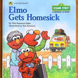 May include: A children's book cover titled "Elmo Gets Homesick" featuring Elmo and family. Elmo is holding a blue stuffed animal, and the family is waving goodbye to a yellow van. The book is part of the Sesame Street series.