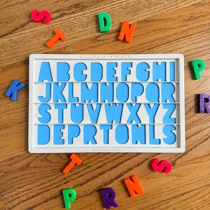 May include: A Fisher-Price alphabet puzzle with blue letter cutouts and a white frame. Scattered around the puzzle are colorful, individual letters in orange, green, pink, purple, and blue. The puzzle is on a wooden surface.