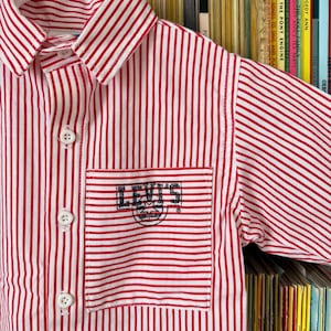 May include: Red and white striped button-up shirt with a pocket featuring the Levi's logo. The shirt has short sleeves and white buttons. The shirt is hanging on a white hanger.