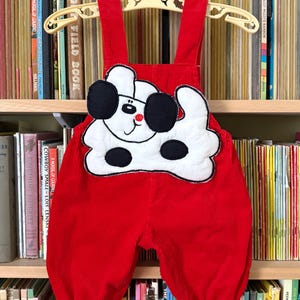 May include: Red corduroy baby overalls with a white and black dog applique. The overalls have adjustable straps and black cuffs. The garment is hanging on a yellow hanger in front of a bookshelf.