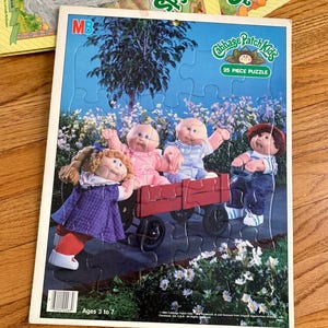 May include: A 25-piece jigsaw puzzle featuring four Cabbage Patch Kids dolls in a red wagon. The dolls are dressed in colorful clothing and are smiling. The puzzle is set against a blue background with white clouds and green trees. The puzzle is labeled "Cabbage Patch Kids 25 Piece Puzzle".