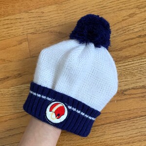 May include: White knit beanie with a blue pom pom and a blue and white striped band. The band has a red and white embroidered football helmet.