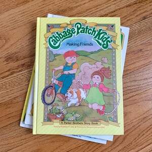May include: A yellow book with the title "Cabbage Patch Kids Making Friends" on the cover. The cover features an illustration of two Cabbage Patch Kids dolls, one riding a bicycle and the other jumping rope. The book is a Parker Brothers story book.