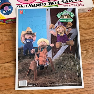 May include: A 25-piece puzzle featuring three Cabbage Patch Kids dolls. The dolls are dressed in different outfits, including a cowboy hat and overalls, a purple dress, and a blue and white dress. The puzzle is set against a background of a blue sky and a barn.