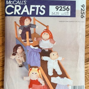 May include: A McCall's Crafts 9256 sewing pattern envelope for 16-inch soft-sculptured dolls. The cover features four dolls in various outfits, including a plaid shirt, a red dress, and a white sleeper, alongside a small wooden toddler gym.