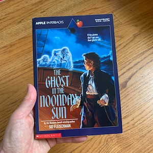 May include: A paperback book cover with a blue background featuring a pirate ship with ghostly figures on the deck. The title of the book is "The Ghost in the Noonday Sun" by Sid Fleischman. The book is published by Scholastic.