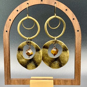 Pode incluir: Um par de brincos em tons de latão com um design único. Cada brinco apresenta um pendente circular grande e texturizado com um círculo interno menor e uma pedra preciosa castanha facetada. Os brincos são exibidos em um suporte de madeira.