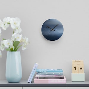 May include: A round wall clock with a blue and white abstract design. The clock has black hands and is mounted on a white wall. A light blue vase with white flowers sits to the left of the clock. Books and a wooden calendar are below.