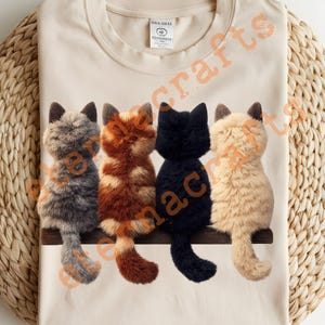 Könnte beinhalten: Cremefarbenes T-Shirt mit einem Design von vier Plüschkatzenfiguren in Grau, Orange, Schwarz und Creme, die auf einem braunen Holzabsatz sitzen. Die Katzen haben flauschiges Fell und sind von hinten zu sehen.