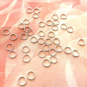 May include: A close-up of a group of silver jump rings, scattered on a pink and white textured surface.